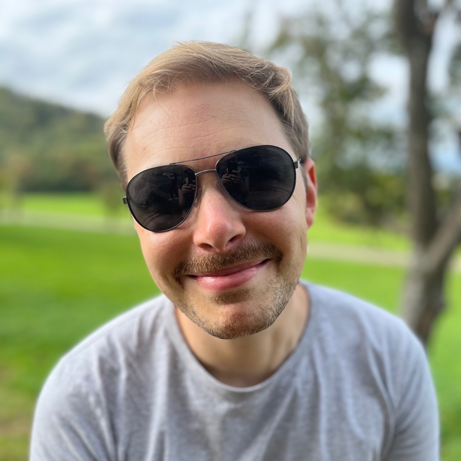 Jacob smirking with sunglasses in front of blurry trees.